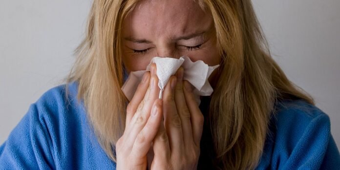 Woman with blonde hair in a blue robe blowing her nose into a tissue held up to her face with eyes closed.