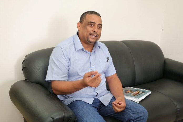 Man in a light blue button-up shirt sits on a black leather sofa, gesturing with his hands as he speaks, with a spiral notebook on the sofa beside him.