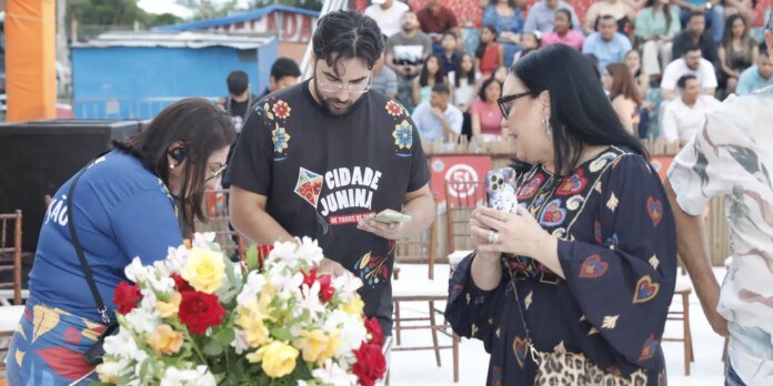 Three people at an outdoor event look at their phones near a colorful flower arrangement, with a crowd in bleachers behind them.