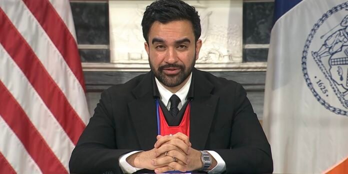 Man in a black suit and tie sits at a desk with hands clasped, wearing a red event lanyard and watch, flanked by the U.S. flag and a seal flag.