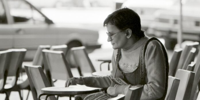 Older woman with short hair and sunglasses sits at an outdoor café table, looking down at the tabletop as her hand rests near the edge of the table.
