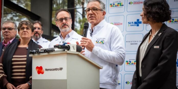 Man in a white lab coat speaks into a microphone at a podium, flanked by colleagues during a press conference with media microphones on display at the front.