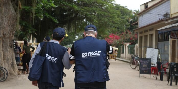 Two IBGE field workers walk away down a dusty street, wearing navy vests with the IBGE logo and bags slung over their shoulders, seen from behind.