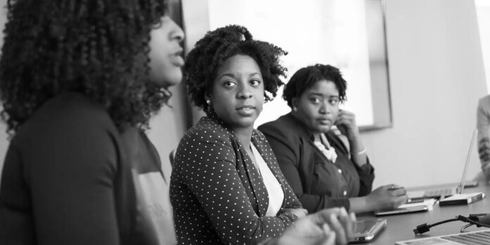 Three women sit at a conference table in a meeting, with the center woman looking toward the camera.