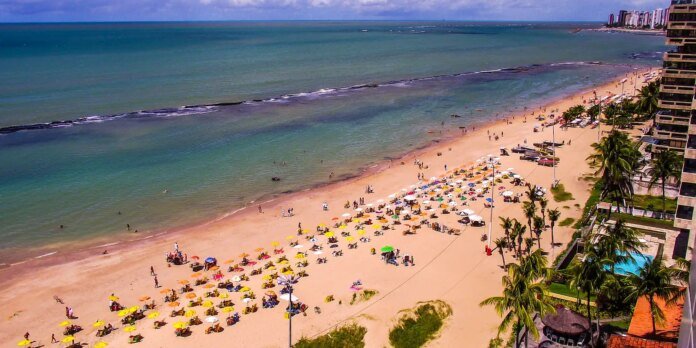 Crowded tropical beach with numerous colorful umbrellas and sunbathers, turquoise sea on the left and high-rise hotels on the right.