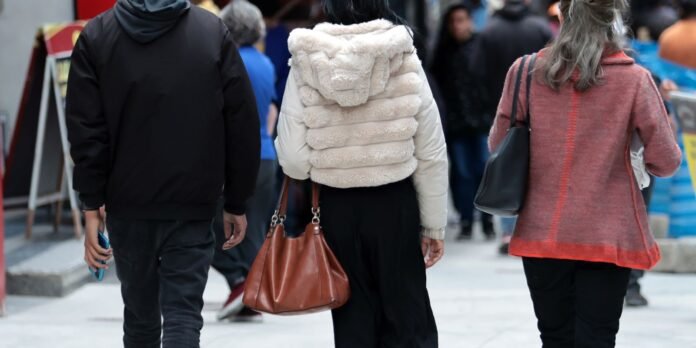 People walking away on a city sidewalk; central figure wears a white faux-fur jacket and carries a brown handbag.