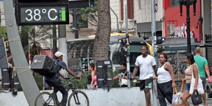 Digital street thermometer showing 38°C in a busy urban scene with pedestrians and a delivery cyclist nearby.