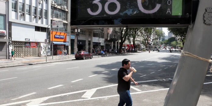 Man in a black shirt walks along a busy city sidewalk, sipping a beverage, with storefronts and a large digital sign overhead.