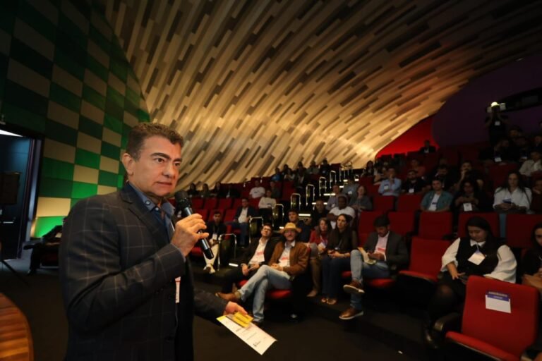 Man in a plaid jacket speaks into a microphone on stage to a seated audience in a modern auditorium with green and tan walls.