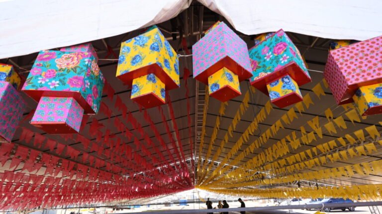 Colorful paper lanterns with floral patterns hang under a white tent, surrounded by red and yellow pennant banners.