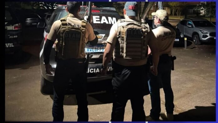 Three armed police officers in tan tactical vests stand with a police SUV at night, facing away from the camera.