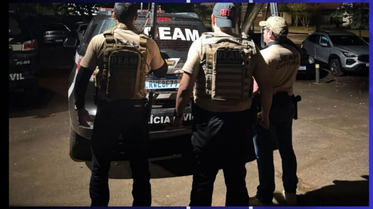 Three armed police officers in tan tactical vests stand with a police SUV at night, facing away from the camera.