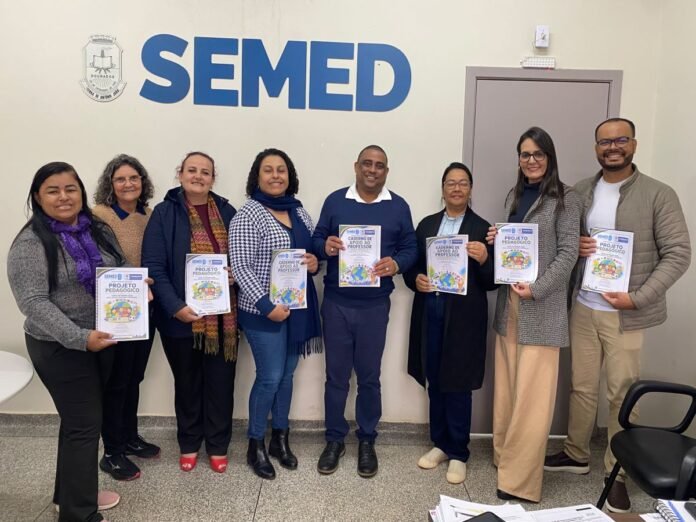Group of nine adults standing in a hallway in front of a SEMED sign, each holding a booklet labeled 'Projeto Pedagógico' (educational project).