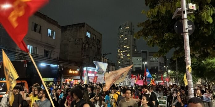 Nighttime street protest with a large crowd carrying colorful banners and flags, city buildings in the background.
