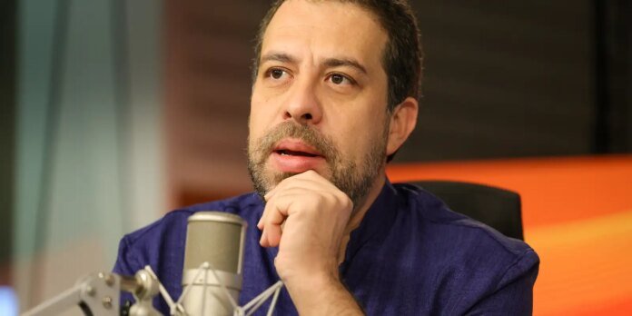 Close-up of a man with a beard sitting at a microphone in a recording studio, looking thoughtful with his hand resting on his chin.