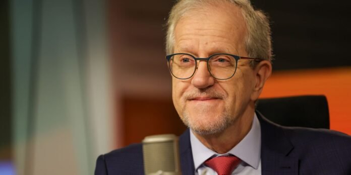 Portrait of an older man with glasses in a navy suit and red tie, speaking into a microphone in a studio.