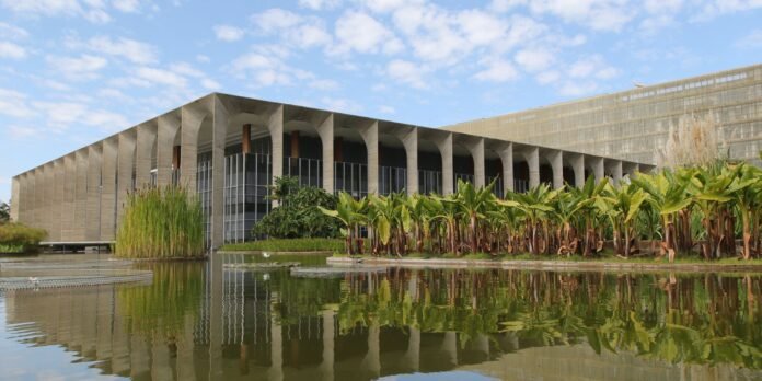 Modern concrete building with an arched colonnade beside a reflective pond and tropical plants along the edge.