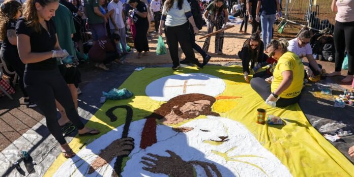 Volunteers painting a large sand mural on a yellow tarp, depicting a bearded man with a halo and a cross in a crowd setting.