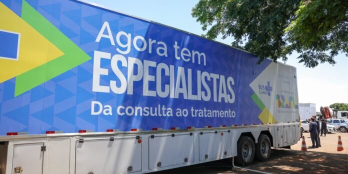 Large blue mobile health trailer with Portuguese slogan Agora tem ESPECIALISTAS Da consulta ao tratamento; people and traffic cones nearby in a parking lot.