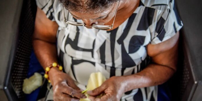 Close-up of an older person's hands weaving a green stem, wearing a black-and-white patterned shirt and a yellow bead bracelet.