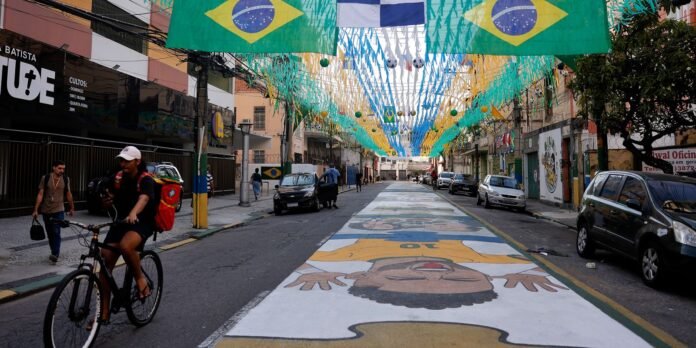 Brazilian street festival: green-yellow flags and blue banners overhead; a cyclist with a red backpack rides past parked cars on a decorated road widening into a mural.
