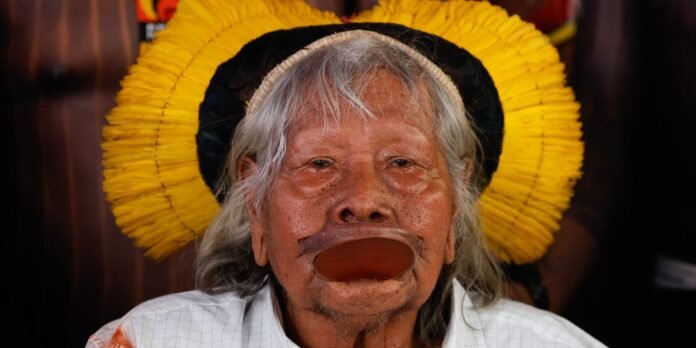 Portrait of an elderly Indigenous woman wearing a large lip plate and a bright yellow circular headdress, in a white checkered shirt