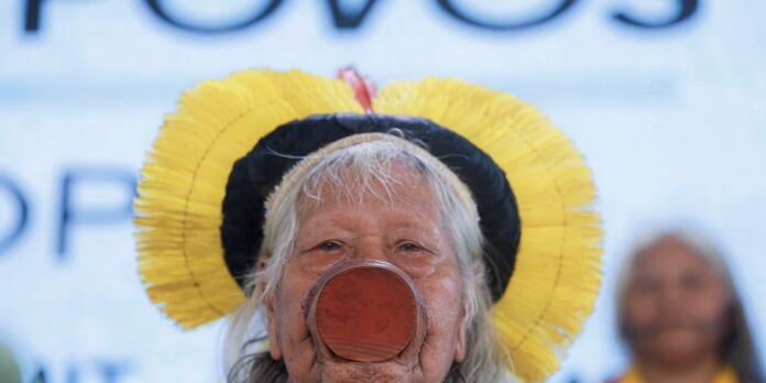 Older woman wearing a large red clown nose and a bright yellow hat, smiling at a festival or event while a blurred figure stands behind her