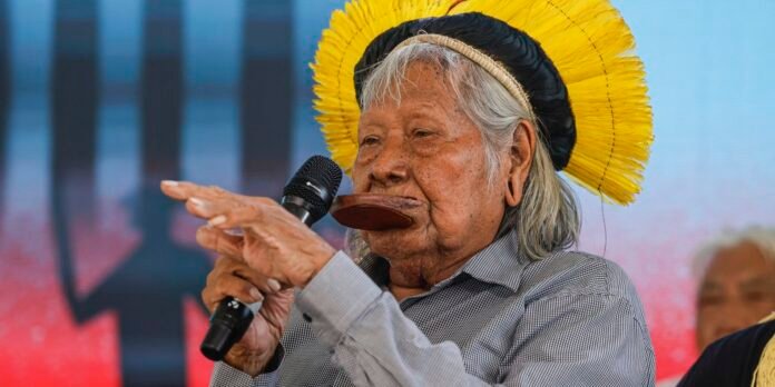 Older person wearing a bright yellow circular headdress and checkered shirt speaks into a handheld microphone on stage.