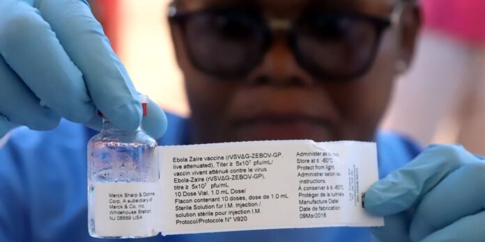 Healthcare worker in blue gloves holds a vial of Ebola vaccine with a printed label, preparing it for use.