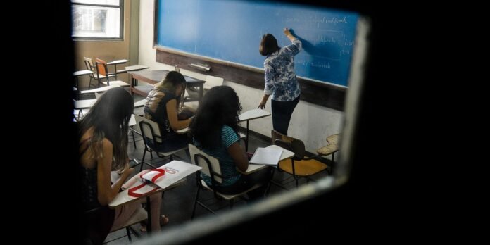Teacher writing on a blue chalkboard at the front of a classroom while students sit at desks with notebooks and bags nearby.