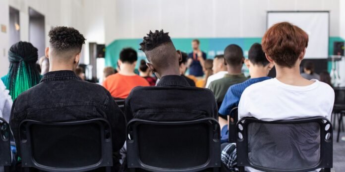 Audience of diverse people seated in a conference room, facing a speaker at the front with a projection screen visible in the background.