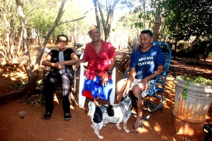Three adults sit outside under trees, smiling, with a small dog in front on a dirt yard.