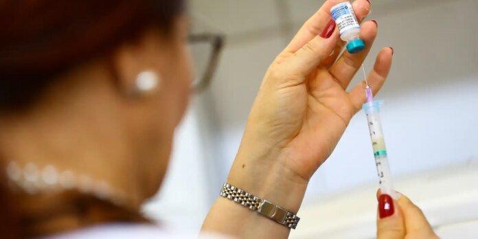A medical professional's hands draw liquid from a vial into a syringe for an injection, with a wristwatch visible on the left hand.