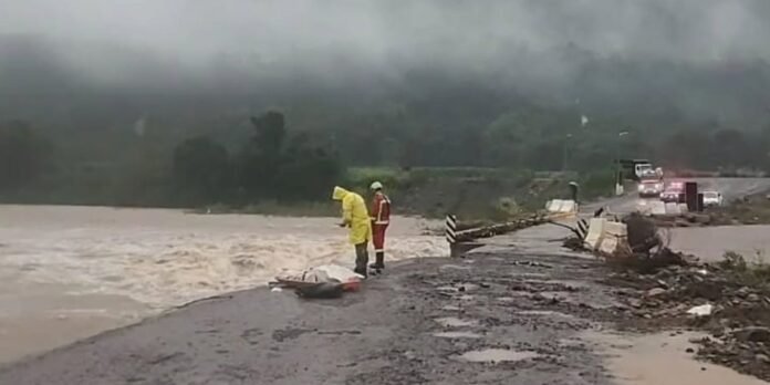 Emergency responders in yellow rain gear stand on a flooded road, surveying debris and water as storm clouds roll in behind a collapsed barrier.
