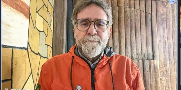 Man with gray beard and round glasses wearing an orange jacket, standing in front of a wooden wall indoors.
