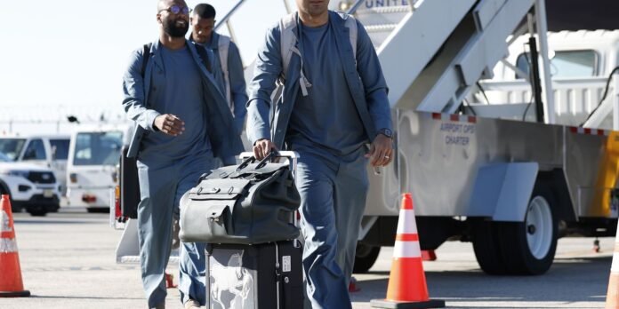 Airline ground crew in blue uniforms walk with a rolling black duffel near a jet bridge and orange traffic cones in the foreground.