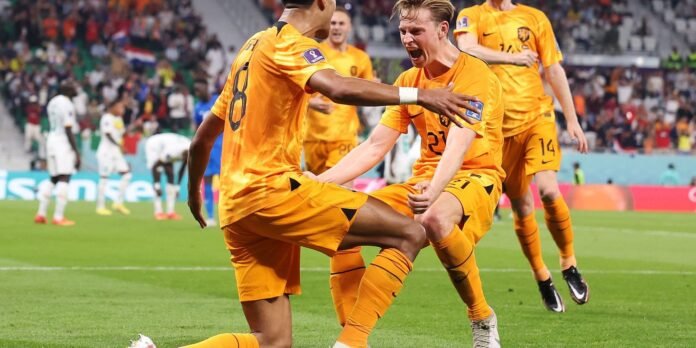 Dutch soccer players in orange celebrate a goal on the field, one kicking the ball while teammates cheer nearby.