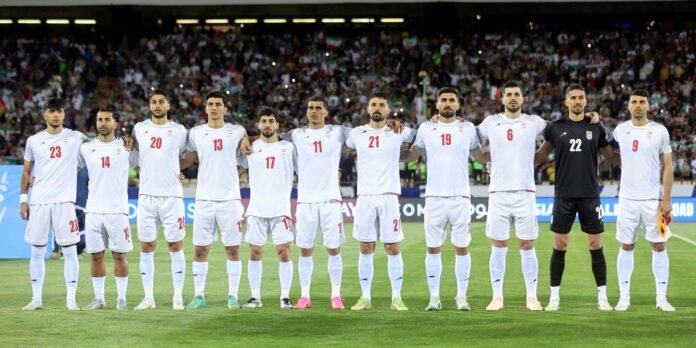 Eleven soccer players in white uniforms with red numbers stand shoulder to shoulder on a grass field before a match, with a crowded stadium in the background.