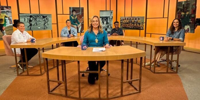 Five people sit around curved wooden desks in a TV studio with orange panels, centered on a smiling host in a turquoise top holding papers.