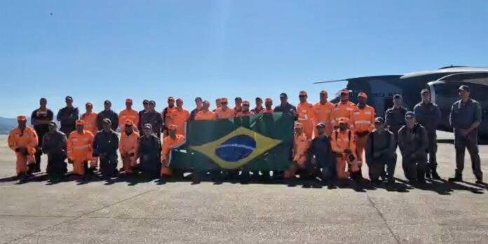 Group of airport workers in orange safety overalls kneeling and standing on a tarmac, holding a Brazilian flag under a clear blue sky.