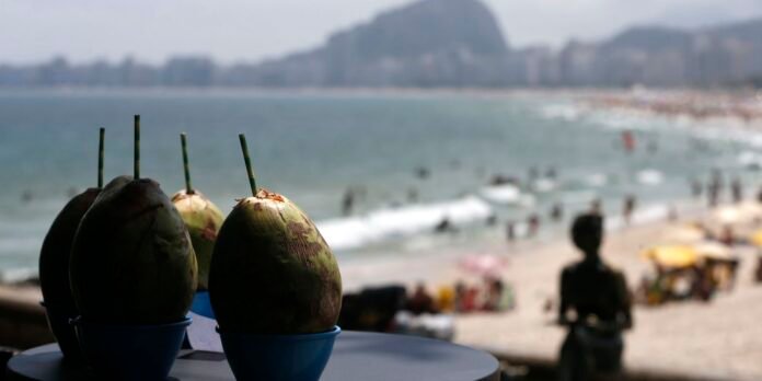Three coconuts with striped green straws in blue cups on a table at a seaside cafe; blurred beach and sea in the background.