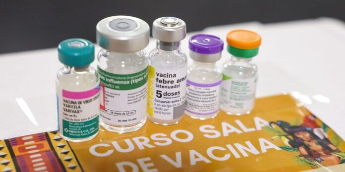 Five small vaccine vials with different colored caps lined up on an orange poster that reads 'CURSO SALA DE VACINA' (vaccine education scene).
