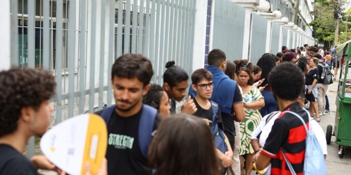 A long queue of diverse people standing along a sidewalk beside a tall grey fence, many wearing backpacks; a green cart is on the curb to the right.