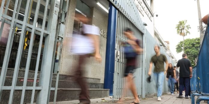 Pedestrians walking along a gated storefront on a city sidewalk, some blurred from motion, with blue metal bars and a palm tree in the background