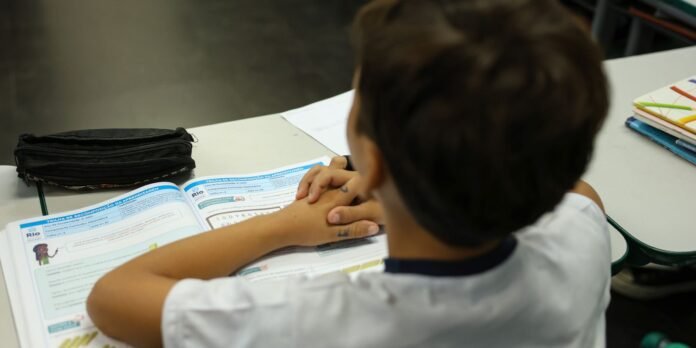 Student sits at a desk with hands folded over an open workbook, a black pencil case beside them.