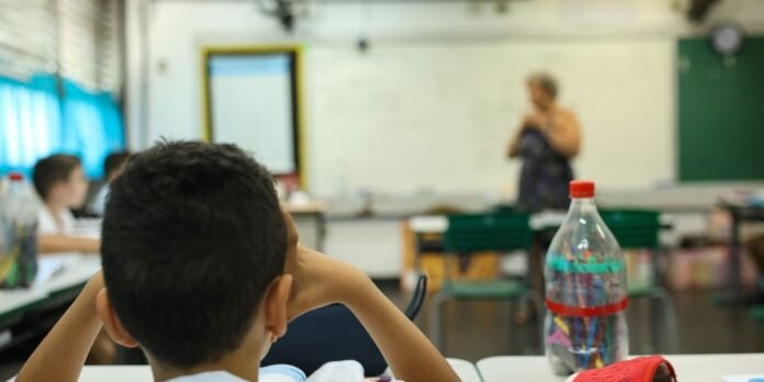 Student sitting at a desk in a classroom, facing a blurred teacher at the whiteboard; a bottle with colored pencils stands on the desk in the foreground.