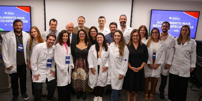 Group of clinicians and researchers in white lab coats posing for a group photo in a conference room, with presentation screens in the background.