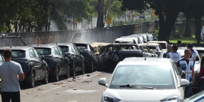 Line of burned-out cars along a street with smoke and bystanders in vests nearby, indicating a vehicle fire aftermath.