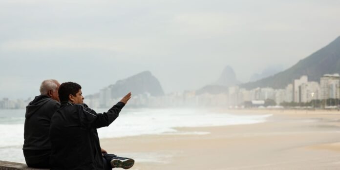 Two elderly people sit on a sandy beach facing the waves, with a city and mountains in the distance; one raises a hand as if pointing.