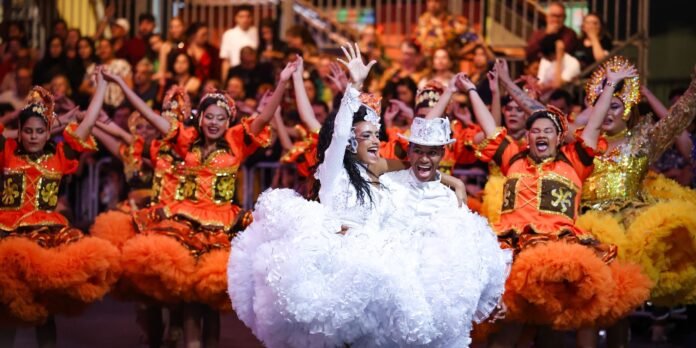 Couple in white outfits dancing center stage among dancers in bright orange costumes at a lively parade or festival.